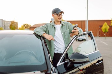 Gözlüklü, telefonu olan yakışıklı bir adam otoparkta arabasının yanında duruyor. Bir arabanın yanında şık bir adam telefonuyla günbatımının tadını çıkarıyor. Ulaşım, eğlence konsepti.