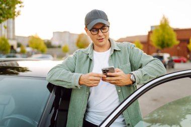 Gözlüklü, telefonu olan yakışıklı bir adam otoparkta arabasının yanında duruyor. Bir arabanın yanında şık bir adam telefonuyla günbatımının tadını çıkarıyor. Ulaşım, eğlence konsepti.