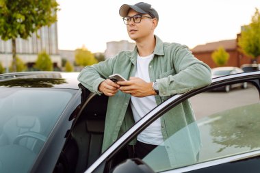 Gözlüklü, telefonu olan yakışıklı bir adam otoparkta arabasının yanında duruyor. Bir arabanın yanında şık bir adam telefonuyla günbatımının tadını çıkarıyor. Ulaşım, eğlence konsepti.