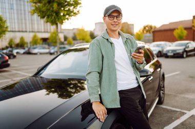 Gözlüklü, telefonu olan yakışıklı bir adam otoparkta arabasının yanında duruyor. Bir arabanın yanında şık bir adam telefonuyla günbatımının tadını çıkarıyor. Ulaşım, eğlence konsepti.