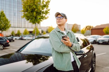 Gözlüklü, telefonu olan yakışıklı bir adam otoparkta arabasının yanında duruyor. Bir arabanın yanında şık bir adam telefonuyla günbatımının tadını çıkarıyor. Ulaşım, eğlence konsepti.