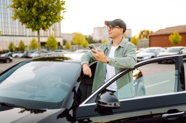 Gözlüklü, telefonu olan yakışıklı bir adam otoparkta arabasının yanında duruyor. Bir arabanın yanında şık bir adam telefonuyla günbatımının tadını çıkarıyor. Ulaşım, eğlence konsepti.