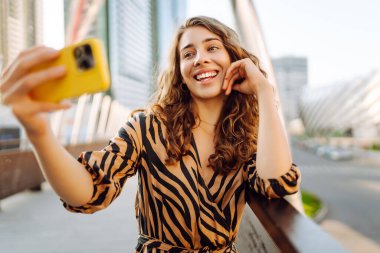 Şık giyinmiş, elinde telefonla Metropolis manzaralı selfie çeken mutlu bir kadın. Genç bir turist blogu yazıp dışarıda eğleniyor. Blogculuk, turizm konsepti.