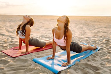 Gün batımı sahili manzaralı minderlerde yoga yapan iki genç kadın. Spor giyimli barışçıl arkadaşlar dengelerini buluyorlar, dışarıda çalışmanın tadını çıkarıyorlar. Etkin yaşam biçimi.