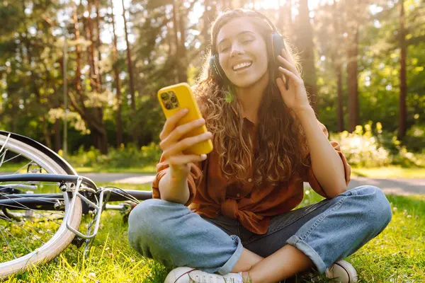 Bisiklet sürdükten sonra çimde oturan kablosuz kulaklıklı genç bir kadın. Güzel bir kadın parkta güneşli bir günün tadını çıkarıyor, müzik dinliyor ve bisiklete biniyor. Etkin yaşam biçimi.