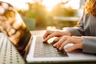 Dizüstü bilgisayarda günbatımı ışınlarıyla yazı yazan genç bir kadının elleri. Dışarıda dizüstü bilgisayarı olan serbest çalışan kadınların yakın çekimleri. İş, uzak çalışma konsepti.