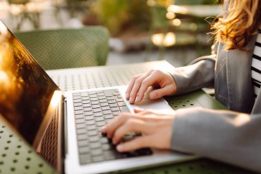 Dizüstü bilgisayarda günbatımı ışınlarıyla yazı yazan genç bir kadının elleri. Dışarıda dizüstü bilgisayarı olan serbest çalışan kadınların yakın çekimleri. İş, uzak çalışma konsepti.