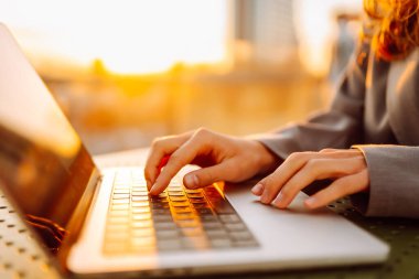 Dizüstü bilgisayarda günbatımı ışınlarıyla yazı yazan genç bir kadının elleri. Dışarıda dizüstü bilgisayarı olan serbest çalışan kadınların yakın çekimleri. İş, uzak çalışma konsepti.