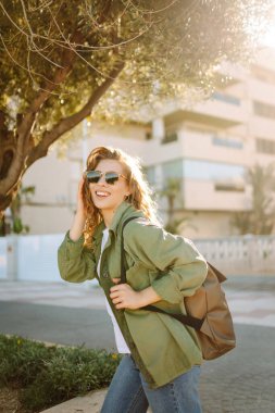 Güneşli şehir caddesinde gün batımında yürüyen güzel bir kadın. Sırt çantalı ve güneş gözlüklü mutlu turist dışarıda havanın tadını çıkarıyor. Hafta sonu ve eğlence konsepti.