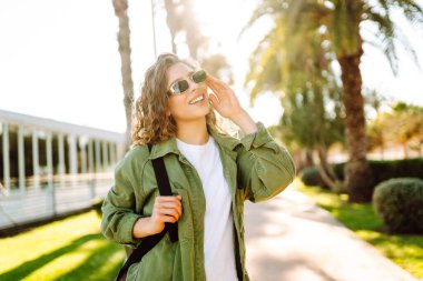 Güneşli şehir caddesinde gün batımında yürüyen güzel bir kadın. Sırt çantalı ve güneş gözlüklü mutlu turist dışarıda havanın tadını çıkarıyor. Hafta sonu ve eğlence konsepti.