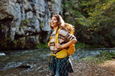 Sırt çantalı genç bir kadın dağ nehri yakınlarındaki dağ manzarasının tadını çıkarıyor. Mutlu kadın gezgin özgürlüğü hisseder, dağlarda yol boyunca yürür. Yürüyüş ve doğa konsepti.