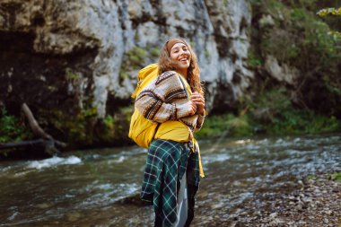 Sırt çantalı genç bir kadın dağ nehri yakınlarındaki dağ manzarasının tadını çıkarıyor. Mutlu kadın gezgin özgürlüğü hisseder, dağlarda yol boyunca yürür. Yürüyüş ve doğa konsepti.