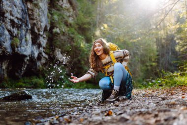 Sırt çantalı genç bir kadın dağ nehri yakınlarındaki dağ manzarasının tadını çıkarıyor. Mutlu kadın gezgin özgürlüğü hisseder, dağlarda yol boyunca yürür. Yürüyüş ve doğa konsepti.