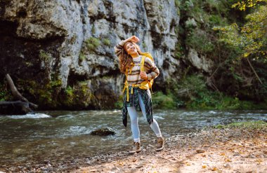Sırt çantalı genç bir kadın dağ nehri yakınlarındaki dağ manzarasının tadını çıkarıyor. Mutlu kadın gezgin özgürlüğü hisseder, dağlarda yol boyunca yürür. Yürüyüş ve doğa konsepti.