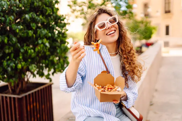 Sokaktaki yemek kutusundan makarna yiyen güzel bir kadının portresi. Gün batımında yemek yiyen genç bir kadın. Fast food konsepti, seyahat.
