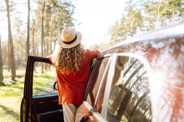 Mutlu genç kadın açık arabanın yanında duruyor ormanda günbatımının tadını çıkarıyor. Arabanın yanındaki şapkalı kadın gezginin arka görüntüsü. Yolculuk konsepti. Etkin yaşam biçimi.