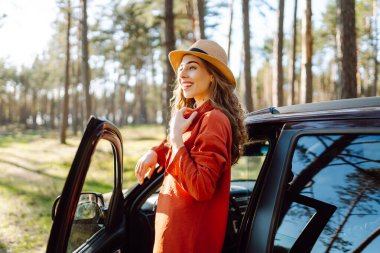 Mutlu genç kadın açık arabanın yanında duruyor ormanda günbatımının tadını çıkarıyor. Arabanın yanındaki şapkalı kadın gezginin arka görüntüsü. Yolculuk konsepti. Etkin yaşam biçimi.