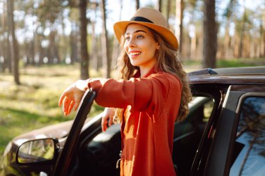 Mutlu genç kadın açık arabanın yanında duruyor ormanda günbatımının tadını çıkarıyor. Arabanın yanındaki şapkalı kadın gezginin arka görüntüsü. Yolculuk konsepti. Etkin yaşam biçimi.