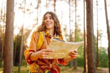 Sırt çantası ve haritası olan genç bir bayan yürüyüşçü yürüyüş parkurunda yön arıyor. Yürüyüş ve günbatımının keyfini çıkaran neşeli bir kadın. Macera konsepti. Etkin yaşam biçimi.