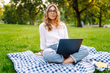 Yeşil parktaki bir çimenlikte dizüstü bilgisayarı ve kablosuz kulaklıkla battaniyenin üzerinde oturan genç bir kadının portresi. Dışarıda kulaklıkla müzik dinlemekten hoşlanan güzel bir bayan serbest yazar. Eğitim kavramı.