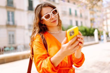 Sunny City caddesinde telefonla yürüyen genç bir kadın. Gülümseyen turist yeni şehrin tadını çıkarıyor ve gün batımında selfie çekiyor. Tatil konsepti, selfie. Etkin yaşam biçimi.