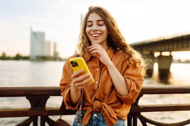 Elinde telefonla gülümseyen kadın setin üzerinde günbatımının tadını çıkarıyor. Güzel bir kadın köprü manzarasına bakarken mesajlaşıyor ve blog yazıyor. Yürüme konsepti. Yaşam biçimi.