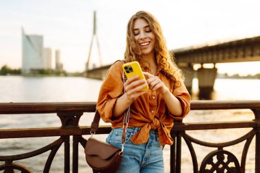 Elinde telefonla gülümseyen kadın setin üzerinde günbatımının tadını çıkarıyor. Güzel bir kadın köprü manzarasına bakarken mesajlaşıyor ve blog yazıyor. Yürüme konsepti. Yaşam biçimi.