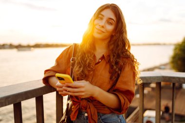 Elinde telefonla gülümseyen kadın setin üzerinde günbatımının tadını çıkarıyor. Güzel bir kadın köprü manzarasına bakarken mesajlaşıyor ve blog yazıyor. Yürüme konsepti. Yaşam biçimi.