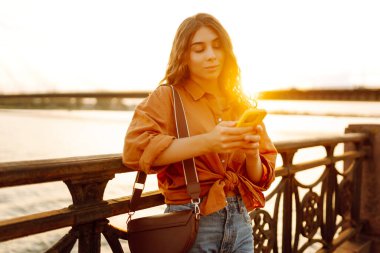 Elinde telefonla gülümseyen kadın setin üzerinde günbatımının tadını çıkarıyor. Güzel bir kadın köprü manzarasına bakarken mesajlaşıyor ve blog yazıyor. Yürüme konsepti. Yaşam biçimi.