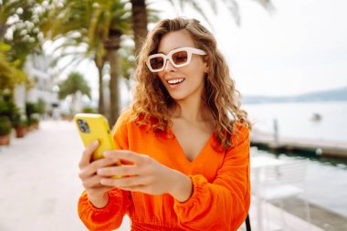 Parlak turuncu elbiseli güzel bir kadının portresi. Telefonu güneşli sette yürüyor. Telefon blogları olan ve dışarı çıkmaktan hoşlanan bir turist. Turizm, blog konsepti.