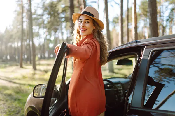 Mutlu genç kadın açık arabanın yanında duruyor ormanda günbatımının tadını çıkarıyor. Arabanın yanındaki şapkalı kadın gezginin arka görüntüsü. Yolculuk konsepti. Etkin yaşam biçimi.