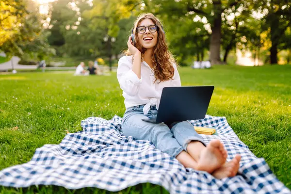 Yeşil parktaki bir çimenlikte dizüstü bilgisayarı ve kablosuz kulaklıkla battaniyenin üzerinde oturan genç bir kadının portresi. Dışarıda kulaklıkla müzik dinlemekten hoşlanan güzel bir bayan serbest yazar. Eğitim kavramı.