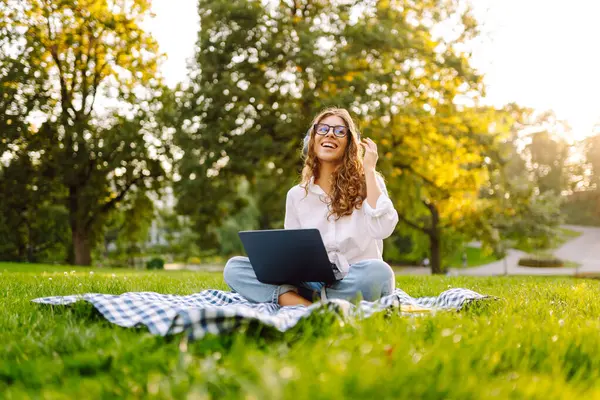 Yeşil parktaki bir çimenlikte dizüstü bilgisayarı ve kablosuz kulaklıkla battaniyenin üzerinde oturan genç bir kadının portresi. Dışarıda kulaklıkla müzik dinlemekten hoşlanan güzel bir bayan serbest yazar. Eğitim kavramı.