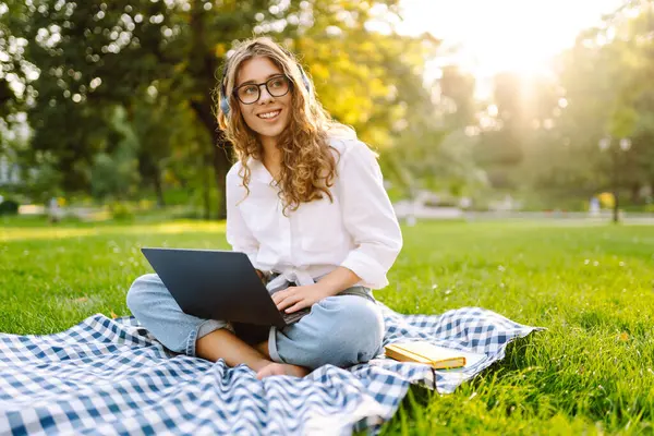 Yeşil parktaki bir çimenlikte dizüstü bilgisayarı ve kablosuz kulaklıkla battaniyenin üzerinde oturan genç bir kadının portresi. Dışarıda kulaklıkla müzik dinlemekten hoşlanan güzel bir bayan serbest yazar. Eğitim kavramı.