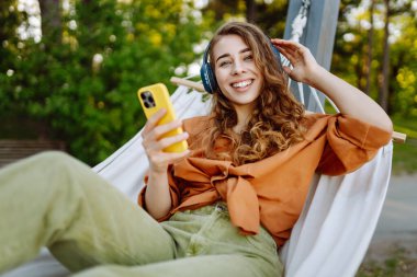 Kablosuz kulaklıklı ve telefonlu şık bir kadın güneşli parkta hamakta uzanıyor. Güzel bir kadın müzik dinlemeyi ya da podcast yapmayı seviyor. Rahatlama ve teknoloji kavramı.