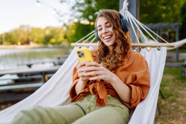 Kablosuz kulaklıklı ve telefonlu şık bir kadın güneşli parkta hamakta uzanıyor. Güzel bir kadın müzik dinlemeyi ya da podcast yapmayı seviyor. Rahatlama ve teknoloji kavramı.