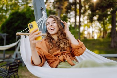Kablosuz kulaklıklı ve telefonlu şık bir kadın güneşli parkta hamakta uzanıyor. Güzel bir kadın müzik dinlemeyi ya da podcast yapmayı seviyor. Rahatlama ve teknoloji kavramı.