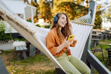 Kablosuz kulaklıklı ve telefonlu şık bir kadın güneşli parkta hamakta uzanıyor. Güzel bir kadın müzik dinlemeyi ya da podcast yapmayı seviyor. Rahatlama ve teknoloji kavramı.