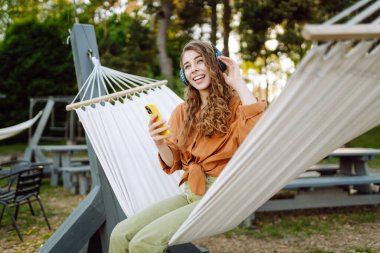 Kablosuz kulaklıklı ve telefonlu şık bir kadın güneşli parkta hamakta uzanıyor. Güzel bir kadın müzik dinlemeyi ya da podcast yapmayı seviyor. Rahatlama ve teknoloji kavramı.