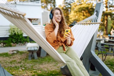 Kablosuz kulaklıklı ve telefonlu şık bir kadın güneşli parkta hamakta uzanıyor. Güzel bir kadın müzik dinlemeyi ya da podcast yapmayı seviyor. Rahatlama ve teknoloji kavramı.