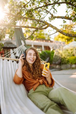 Kablosuz kulaklıklı ve telefonlu şık bir kadın güneşli parkta hamakta uzanıyor. Güzel bir kadın müzik dinlemeyi ya da podcast yapmayı seviyor. Rahatlama ve teknoloji kavramı.