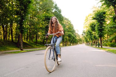 Yeşil bir parkta bisiklet süren genç bir kadın. Neşeli kadın hafta sonunun tadını çıkarıyor, gün batımında bisiklet sürüyor. Aktif yaşam tarzı. Joy, hafta sonu konsepti.