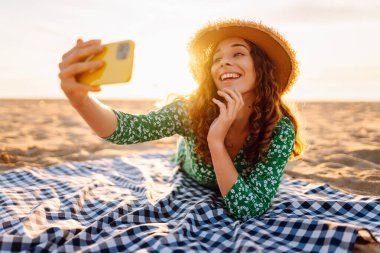 Hasır şapkalı ve parlak elbiseli neşeli bir kadın deniz kenarındaki bir paspasın üzerinde yatarken telefonunda selfie çekiyor. Güzel bir turist deniz manzarası ve bloglardan hoşlanır. Bloglama, hafta sonu konsepti.
