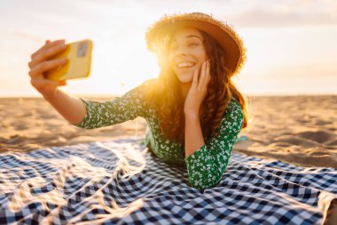 Hasır şapkalı ve parlak elbiseli neşeli bir kadın deniz kenarındaki bir paspasın üzerinde yatarken telefonunda selfie çekiyor. Güzel bir turist deniz manzarası ve bloglardan hoşlanır. Bloglama, hafta sonu konsepti.