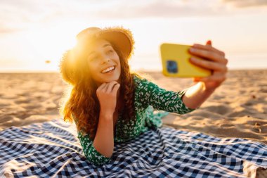 Hasır şapkalı ve parlak elbiseli neşeli bir kadın deniz kenarındaki bir paspasın üzerinde yatarken telefonunda selfie çekiyor. Güzel bir turist deniz manzarası ve bloglardan hoşlanır. Bloglama, hafta sonu konsepti.