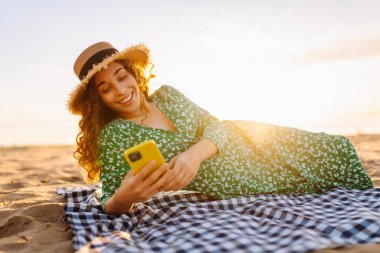 Hasır şapkalı ve parlak elbiseli neşeli bir kadın deniz kenarındaki bir paspasın üzerinde yatarken telefonunda selfie çekiyor. Güzel bir turist deniz manzarası ve bloglardan hoşlanır. Bloglama, hafta sonu konsepti.