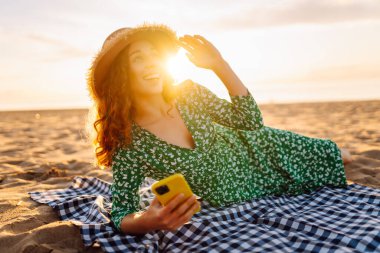 Hasır şapkalı ve parlak elbiseli neşeli bir kadın deniz kenarındaki bir paspasın üzerinde yatarken telefonunda selfie çekiyor. Güzel bir turist deniz manzarası ve bloglardan hoşlanır. Bloglama, hafta sonu konsepti.