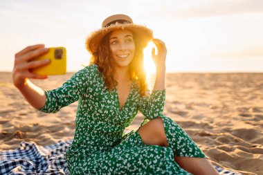 Hasır şapkalı ve parlak elbiseli neşeli bir kadın deniz kenarındaki bir paspasın üzerinde yatarken telefonunda selfie çekiyor. Güzel bir turist deniz manzarası ve bloglardan hoşlanır. Bloglama, hafta sonu konsepti.