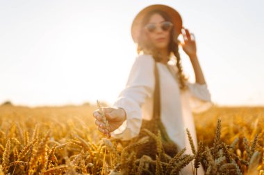 Gün batımında altın buğday kulağına dokunan şapkalı genç bir kadının yakın çekimi. Güzel bir kadın yürümekten zevk alıyor, açık havada özgürlüğü hissediyor. Güzellik ve yürüyüş konsepti.