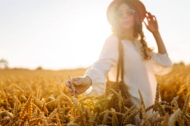 Gün batımında altın buğday kulağına dokunan şapkalı genç bir kadının yakın çekimi. Güzel bir kadın yürümekten zevk alıyor, açık havada özgürlüğü hissediyor. Güzellik ve yürüyüş konsepti.